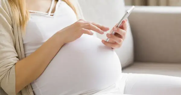 Kobieta w 34. tygodniu ciąży przegląda coś na telefonie, siedząc wygodnie na kanapie.